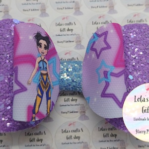May include: A handmade hair bow featuring a cartoon character design. The bow has a purple glitter base with a light blue glitter accent. The center features a cartoon character with star accents. The bow is from Lola's Crafts & Gift Shop.