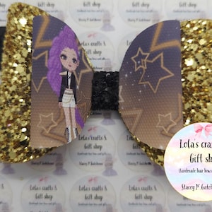 May include: A handmade hair bow with a glittery gold and black design. The bow features a cartoon character with purple hair and a black jacket. The bow is decorated with star patterns and the text "Lola's crafts & gift shop".