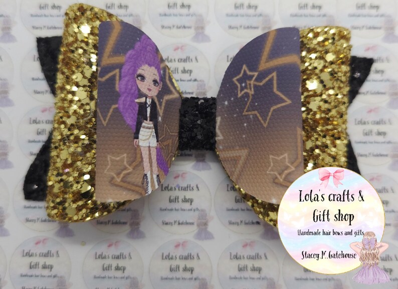 May include: A handmade hair bow with a glittery gold and black design. The bow features a cartoon character with purple hair and a black jacket. The bow is decorated with star patterns and the text "Lola's crafts & gift shop".