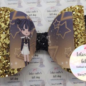 May include: A handmade hair bow featuring a cartoon character with a star and moon design. The bow has gold glitter accents and black details. The bow is from Lola's Crafts & Gift Shop.