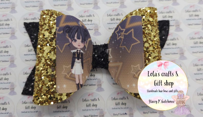 May include: A handmade hair bow featuring a cartoon character with a star and moon design. The bow has gold glitter accents and black details. The bow is from Lola's Crafts & Gift Shop.