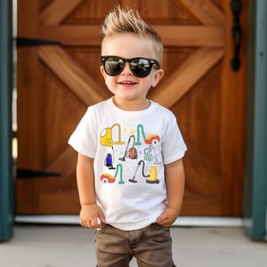 以下が含まれることがあります： 掃除用具、掃除機、ほうき、ちりとりなどがカラフルにデザインされた白いTシャツ。金髪の男の子がサングラスをかけて着ています。