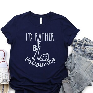以下が含まれることがあります： 白い文字で「I'd rather be vacuuming」と書かれたネイビーブルーのTシャツ。テキストには掃除機のグラフィックが添えられています。