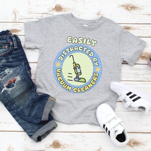 May include: A gray t-shirt with the text "EASILY DISTRACTED BY VACUUM CLEANERS" in a circular design featuring a cartoon vacuum cleaner. The shirt is paired with blue jeans and white sneakers with black stripes.