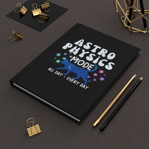 Könnte beinhalten: Schwarzes Notizbuch mit dem Text "Astro Physics Mode" in Weiß, mit einer blauen Katzensilhouette, die auf einer Mondsichel und Sternen geht. Der Text "All Day - Every Day" befindet sich unter der Katze.