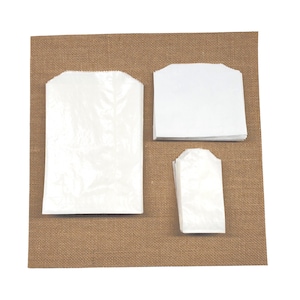 May include: Three white paper bags of different sizes on a brown hessian background. The bags are all made of a thin, crinkly paper and have a simple, classic design.