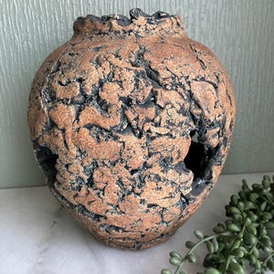 May include: A brown ceramic vase with a textured, cracked surface and several holes. The vase is sitting on a white surface with green plants in the foreground.