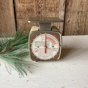 Könnte beinhalten: Eine vintage weiße und braune Zephyr Modell Z-5 Briefwaage mit einem roten Zeiger und einem Zifferblatt, das bis zu 5 Pfund anzeigt. Die Waage hat eine silberne Plattform und eine Tabelle zur Berechnung der Portokosten.