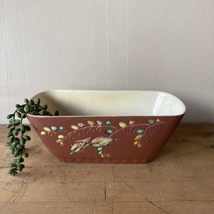 Puede incluir: Un macetero de cerámica marrón con interior blanco y un diseño floral en tonos verdes, amarillos y naranjas.