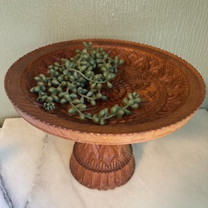 Peut inclure: Un bol sur piédestal en bois sculpté marron avec un bord et une base décoratifs. Le bol contient une branche de plantes succulentes artificielles vertes. Le bol sur piédestal est un objet décoratif.