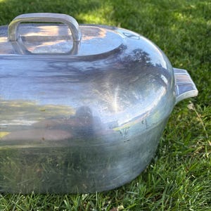 May include: A silver-colored oval metal roasting pan with a matching lid. The lid features a central arched handle with ribbed detailing and a side handle with similar ribbed accents. The surface of the metal is smooth and reflective.