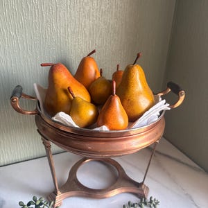 Peut inclure: Un plat de service cuivré contient plusieurs poires artificielles. Les poires sont de différentes tailles et nuances d'orange et de jaune. Le plat a un support décoratif et des poignées noires. Une branche de feuillage vert est au premier plan.