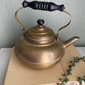 May include: A vintage brass teapot with a black wooden handle and a small spout. The teapot sits on a closed book with a light tan cover. The handle is curved and attached to the body of the teapot with brass fixtures.