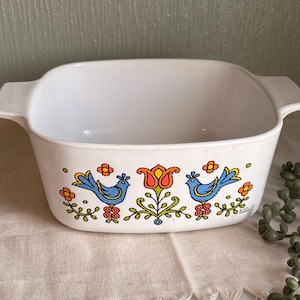 May include: White square casserole dish with handles, decorated with blue birds, orange flowers, and a central orange and yellow floral design. The dish is on a beige fabric.