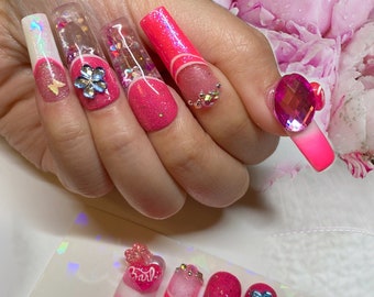 Peut inclure: Ongles longs en acrylique rose et blanc avec des paillettes, des strass et des accents de fleurs et de papillons 3D. Certains ongles ont une base transparente avec des paillettes et des strass.