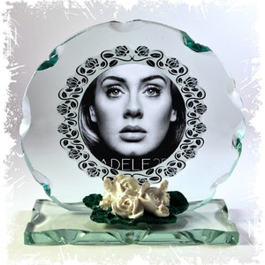 Puede incluir: Un premio de vidrio transparente con una imagen en blanco y negro de Adele, rodeada por un diseño floral. La base del premio tiene un pequeño grupo de rosas blancas con hojas verdes. El texto "ADELE 25" es visible.