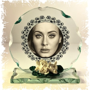 Puede incluir: Un marco de fotos de vidrio transparente con un retrato de una mujer, rodeado por un diseño floral negro. El marco se apoya en una base de vidrio transparente, con rosas blancas y hojas verdes. La imagen tiene un tono sepia.