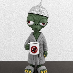 Könnte beinhalten: Eine grüne Alien-Figur mit großen roten Augen, die einen Alufolienhut, einen grauen Bademantel und graue Hausschuhe trägt. Das Alien hält eine weiße Tasse mit einem roten Kreis und einem diagonalen Strich über einer schwarzen Vogelsilhouette.