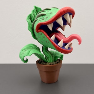 Puede incluir: Una figura de planta carnívora verde con la boca abierta bordeada de rojo, dientes blancos afilados y una larga lengua rosa. La planta tiene hojas texturizadas y está plantada en una pequeña maceta marrón con textura de tierra.