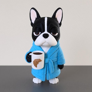Può includere: Una statuetta di un bulldog francese bianco e nero con un'espressione assonnata, che indossa un accappatoio blu brillante. Il cane tiene in mano una tazza bianca con il disegno di un croissant marrone e del liquido scuro.