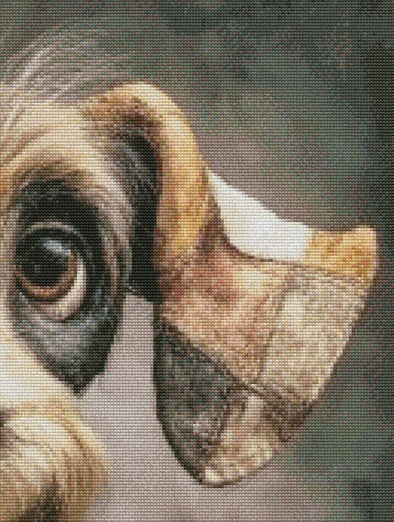 May include: Close-up of a cross-stitch portrait of a dog's eye and ear. The eye is dark brown with a black patch around it. The ear is tan, brown, and white, with a textured appearance.