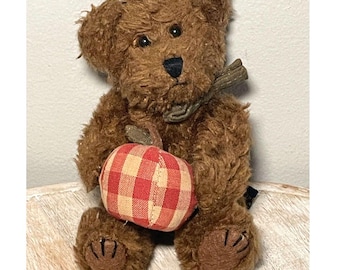 Puede incluir: Un oso de peluche marrón con pelaje texturizado, ojos oscuros y nariz negra. El oso sostiene una manzana de tela rellena con un patrón de cuadros rojos y beige. Una pequeña hoja de tela verde está unida a la parte superior de la manzana.