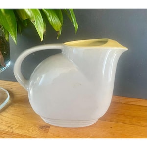 May include: A vintage, off-white ceramic pitcher with a rounded body and a curved handle. The rim of the pitcher is a pale yellow. The pitcher is sitting on a wooden surface.