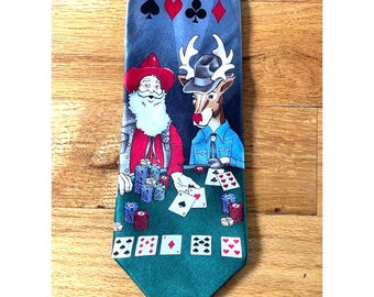 Puede incluir: Una corbata azul con un estampado gráfico de Santa Claus y un reno con sombreros de vaquero, jugando al póquer en una mesa verde con fichas y cartas. La parte superior de la corbata muestra símbolos de palos de cartas.