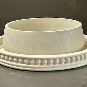 May include: A light-colored, round planter with a matching saucer. The planter has a cylindrical shape and sits inside the saucer. The saucer has a decorative beaded edge. The set is made of a matte, light-colored material.