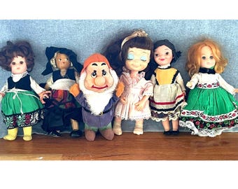 Puede incluir: Una colección de seis muñecas de pie en fila. El grupo incluye cuatro muñecas con vestidos coloridos con detalles florales, una muñeca con un atuendo oscuro y un personaje de peluche con gorro rojo, barba blanca y ropa morada.
