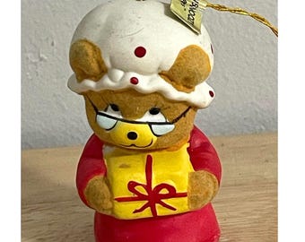 Puede incluir: Un adorno de oso marrón con un gorro blanco de lunares rojos y gafas de montura blanca. El oso viste un traje rojo y sostiene un regalo amarillo con un lazo rojo. Tiene un hilo dorado con una etiqueta que dice SOFTN WOOL en la parte superior.