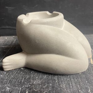 May include: A gray concrete ashtray shaped like a person sitting with their legs crossed. The ashtray has a round opening on top for holding cigarette butts.