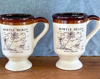 Puede incluir: Dos tazas de cerámica de color crema con bordes y asas marrones. Cada taza presenta una ilustración gráfica marrón de una palmera, un velero y un pez, con el texto MYRTLE BEACH y SOUTH CAROLINA impreso en la parte frontal de las tazas.