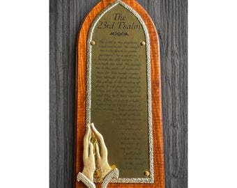 Puede incluir: Una placa de pared de madera con arco apuntado que presenta una placa de metal dorado grabada con el texto del Salmo 23. La placa está decorada con una escultura tridimensional de color crema de dos manos unidas en oración.