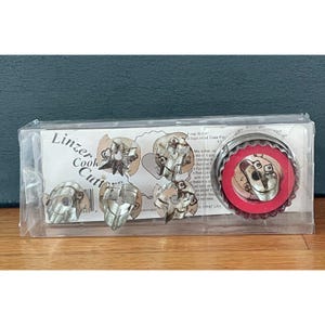 May include: A clear plastic box containing a set of metal Linzer cookie cutters. The set includes one large round scalloped cutter with a red center insert and four smaller interchangeable metal shapes, all displayed on a white backing card.