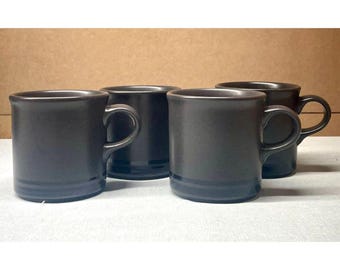 Puede incluir: Un juego de cuatro tazas de cerámica de color marrón oscuro con un acabado mate suave. Cada taza presenta un cuerpo cilíndrico, un asa lateral resistente y un sutil detalle de banda horizontal cerca de la base. Las tazas están dispuestas en una formación de dos por dos.