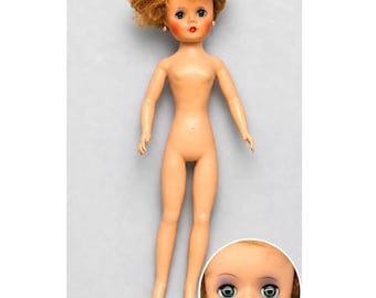 Puede incluir: Una muñeca de piel clara, cabello corto rizado de color jengibre y ojos azules. La muñeca tiene pequeños pendientes de botón blancos, labios pintados de rojo y una expresión facial neutra. Un recuadro circular muestra el rostro en detalle.