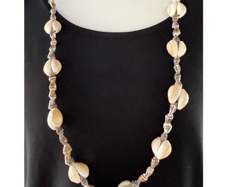 Puede incluir: Un collar largo que presenta una serie de conchas de color blanco roto, lisas y con forma de corazón, que se alternan con cuentas más pequeñas, texturizadas y de tonos metálicos. Las conchas están dispuestas en pares a lo largo.