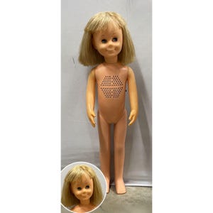 May include: A plastic doll with blonde hair, blue eyes, and a smiling expression. The doll features a light peach skin tone and a hexagonal speaker grille integrated into the chest area. A circular inset shows a close-up view of the doll's face.