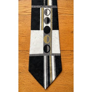 May include: A necktie featuring a geometric pattern with black, white, and gold sections. The design includes a central vertical column displaying a series of circular moon phase icons in black, grey, and gold, set against a textured fabric background.