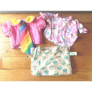 May include: Three doll garments including a pink grid-patterned shirt with a yellow collar and a rainbow-striped tie, a long-sleeved pink shirt with a multi-colored abstract print, and a beige shirt featuring a repeating pattern of small green bears.