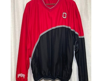 Puede incluir: Una chaqueta cortavientos de manga larga roja y negra con cuello en V. La parte delantera presenta un logotipo de bloque O blanco y rojo en el pecho y el texto Ohio State bordado en blanco en el puño de la manga inferior derecha.