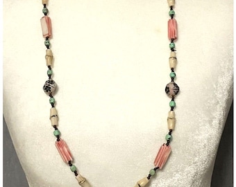 Puede incluir: Un collar largo de cuentas con una alternancia de cuentas cilíndricas color crema, cuentas rosas estriadas y cuentas redondas con patrones florales negros. Pequeñas cuentas turquesas y diminutos espaciadores marrones unen las piezas.
