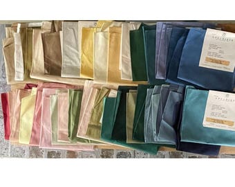 Puede incluir: Una colección de muestras de tela plegadas en un espectro de colores que incluye beige, amarillo, verde y azul. Cada muestra presenta una etiqueta blanca con el texto GP&J BAKER, detallando la composición y el origen.