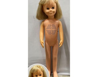 Puede incluir: Una muñeca de plástico con cabello rubio y ojos azules, que presenta una rejilla de altavoz hexagonal en el pecho. La muñeca tiene un acabado liso y de tono claro y está de pie. Un recuadro circular muestra un primer plano de la cara y el pelo.