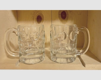Puede incluir: Dos jarras de cerveza de cristal transparente con una superficie exterior texturizada con hoyuelos, una base gruesa y un asa lateral resistente. Las jarras son transparentes y de forma cilíndrica, diseñadas para contener bebidas.