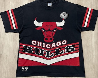 Puede incluir: Una camiseta negra de manga corta con el logotipo de los Chicago Bulls en rojo, blanco y negro. La prenda muestra el texto "CHICAGO BULLS" y "EASTERN CONFERENCE" con rayas rojas y blancas en las mangas y un patrón de chevrón.
