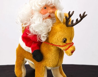 Puede incluir: Una figura de peluche de Santa Claus con barba blanca y traje rojo montado sobre un reno de peluche color canela. El reno tiene astas de color marrón oscuro, un arnés de cuerda roja y una nariz roja. Santa lleva un gorro rojo.