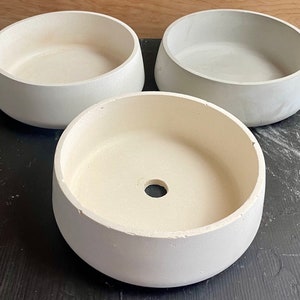 May include: Three white ceramic planters, two with a wide opening and one with a smaller opening in the center. The planters are on a black surface.