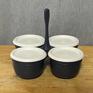 May include: A black serving caddy featuring four individual round containers with white lids. The caddy has a central vertical handle for carrying. Each container is matte black with a smooth finish, and the white lids have a circular ridged design.